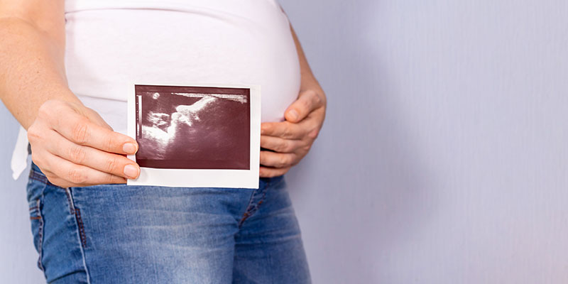 A pregnant woman in jeans and a white T-shirt holds a an ultrasound photo of her baby in her hand on light blue background.
